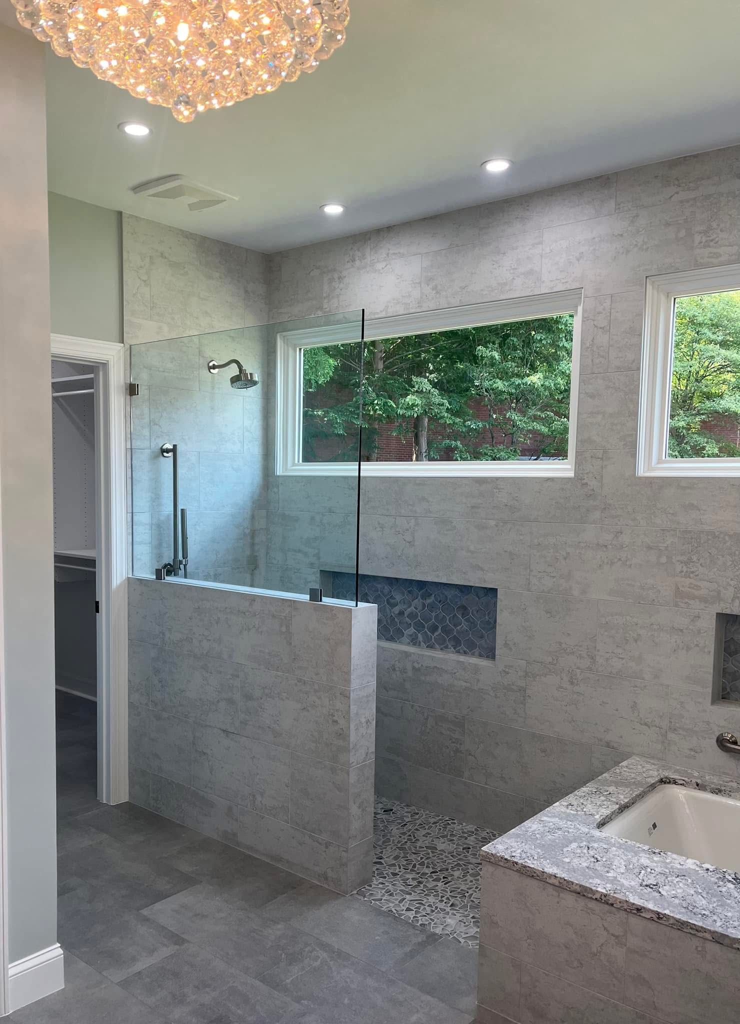 Modern bathroom with gray tiled walls, walk-in shower, and a large window overlooking trees.