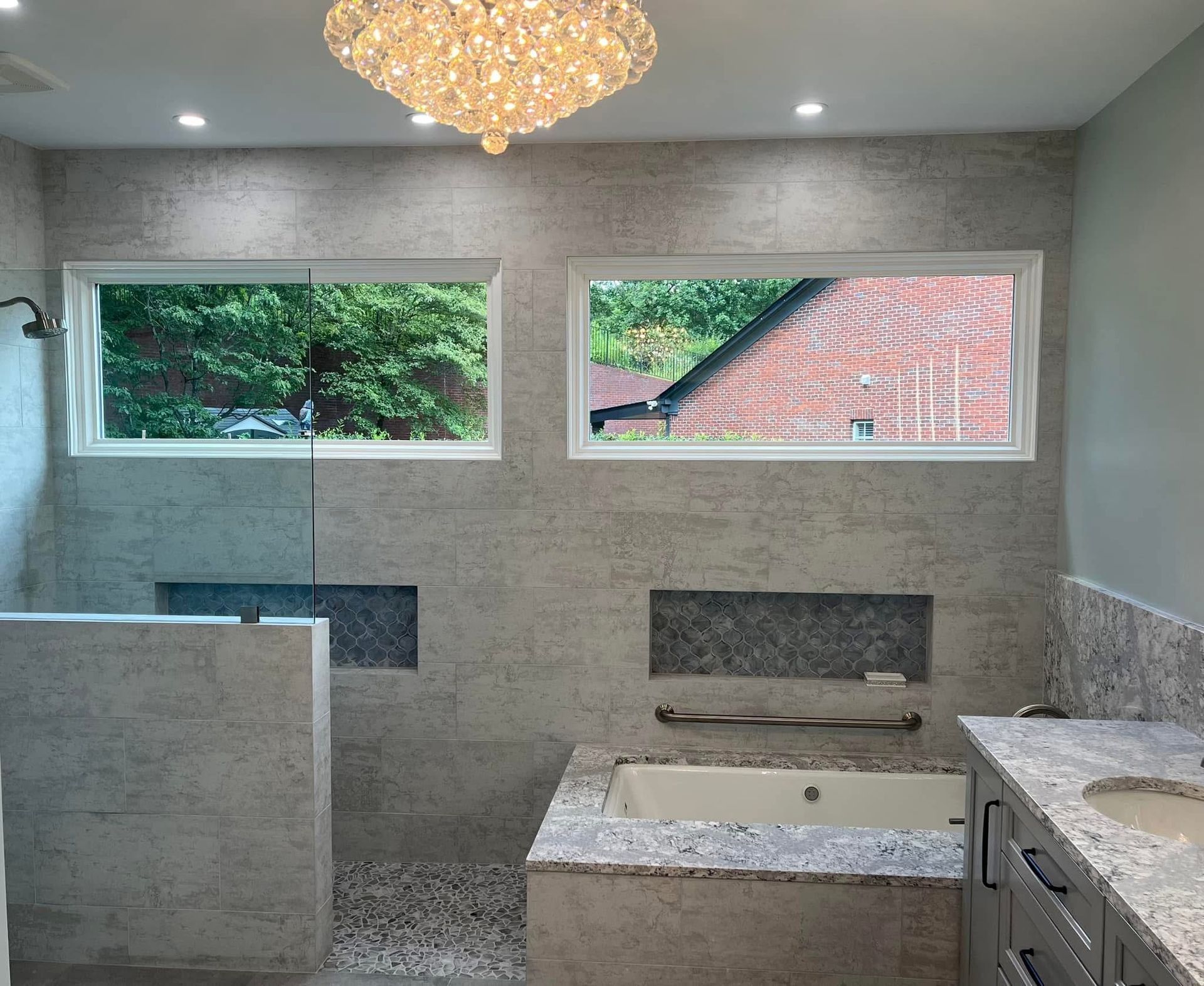 Bathroom with light-colored tile walls, windows, recessed shelving, bathtub, shower, and chandelier.