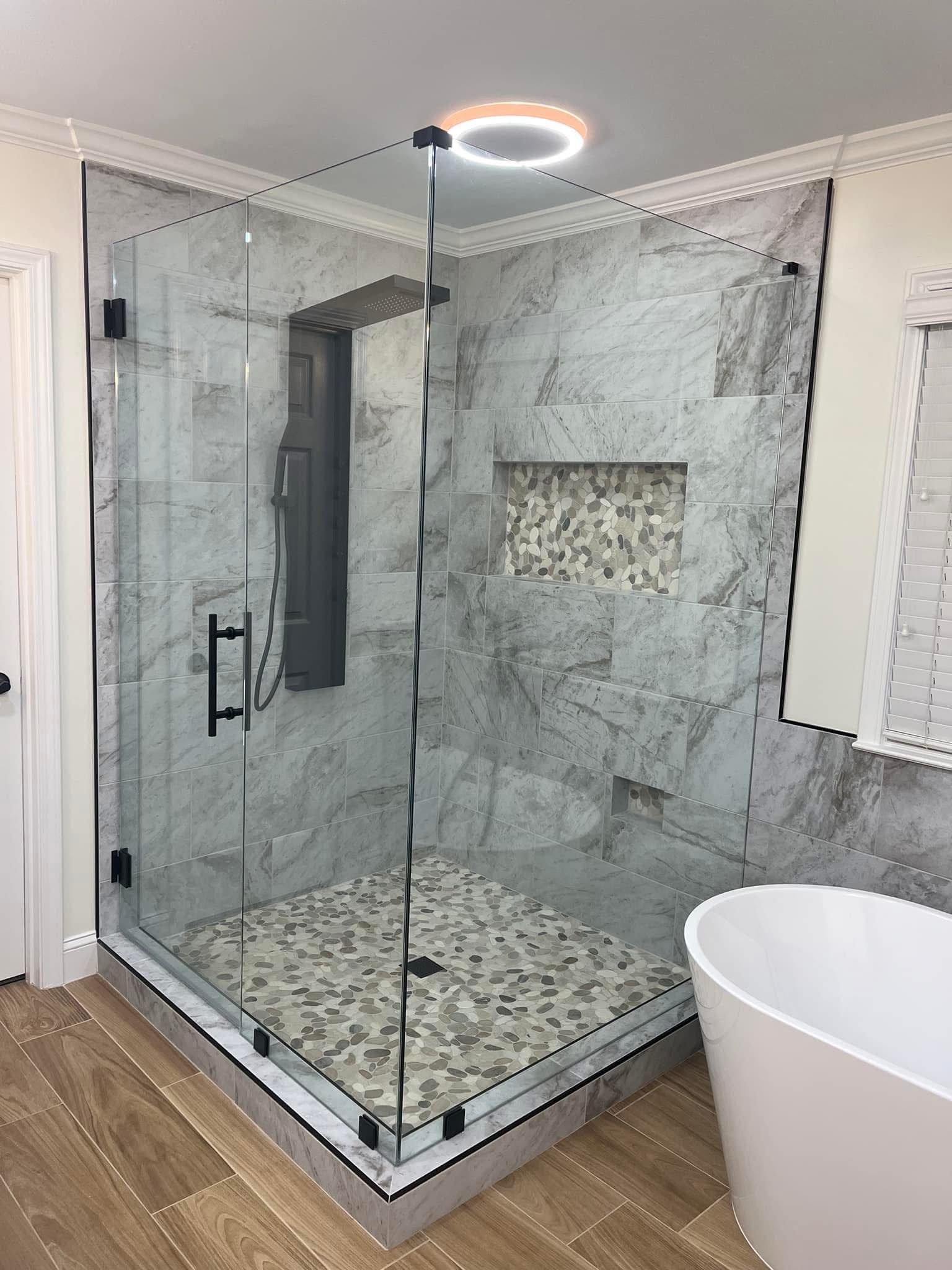 Corner glass shower with stone tile walls, pebble floor, and white tub.