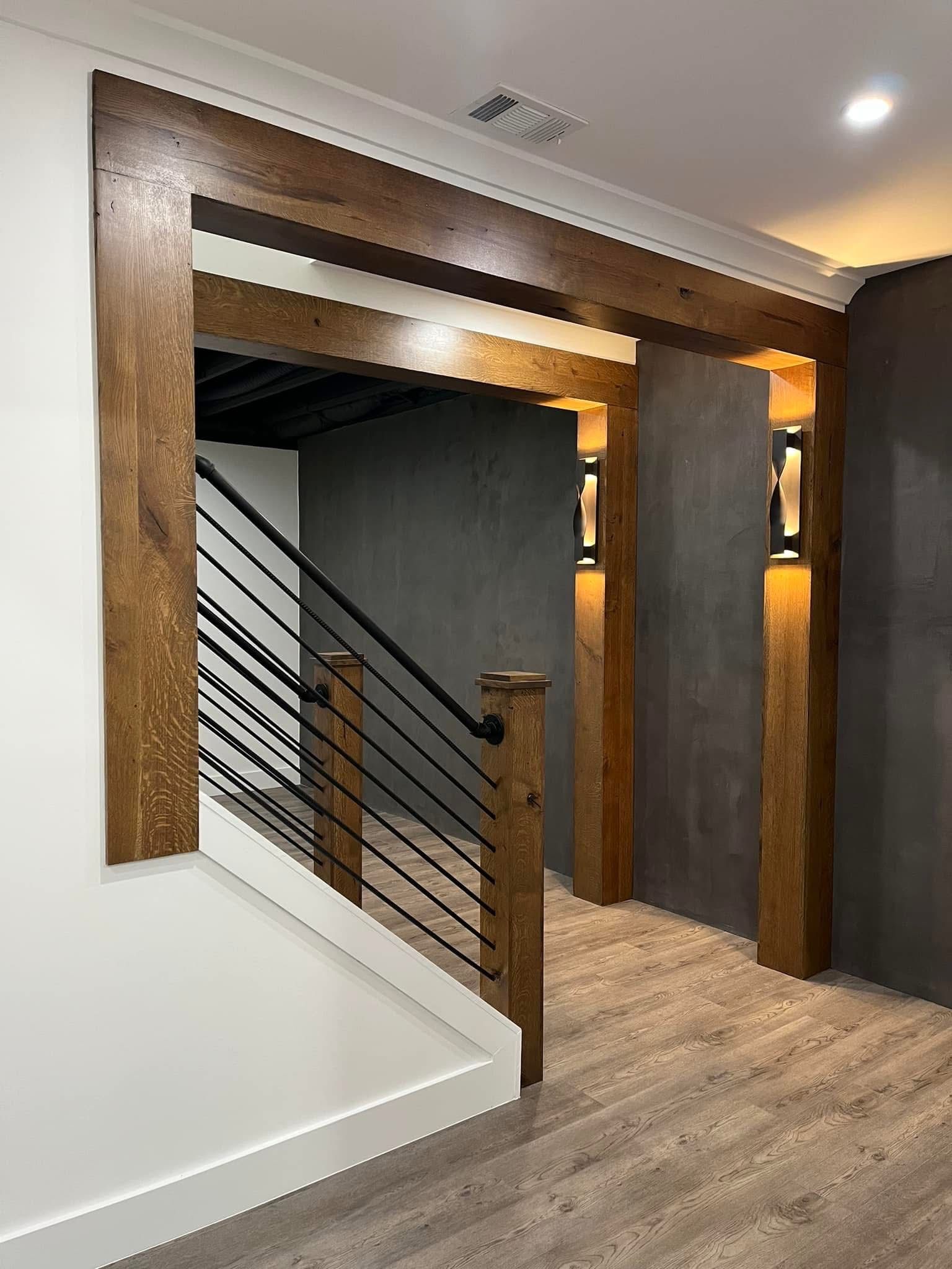 Wooden beams frame a doorway leading to a staircase with black metal railings. Light fixtures are on the wall.