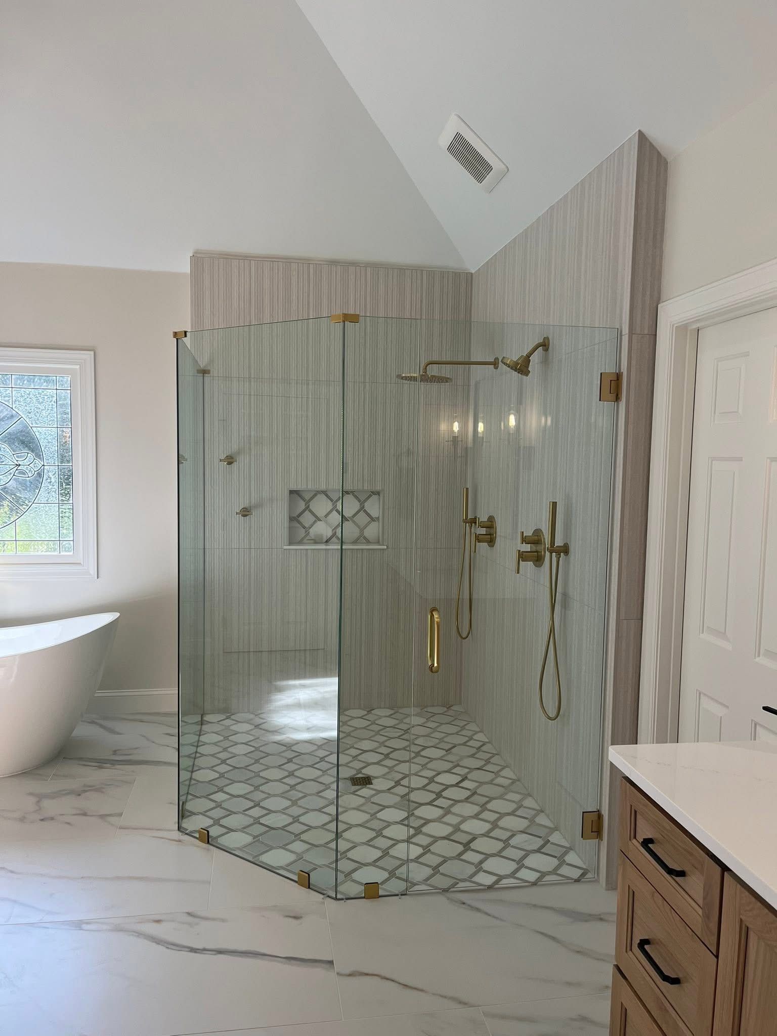 Spacious bathroom with glass shower, patterned floor, and white walls. Gold fixtures and a large window.