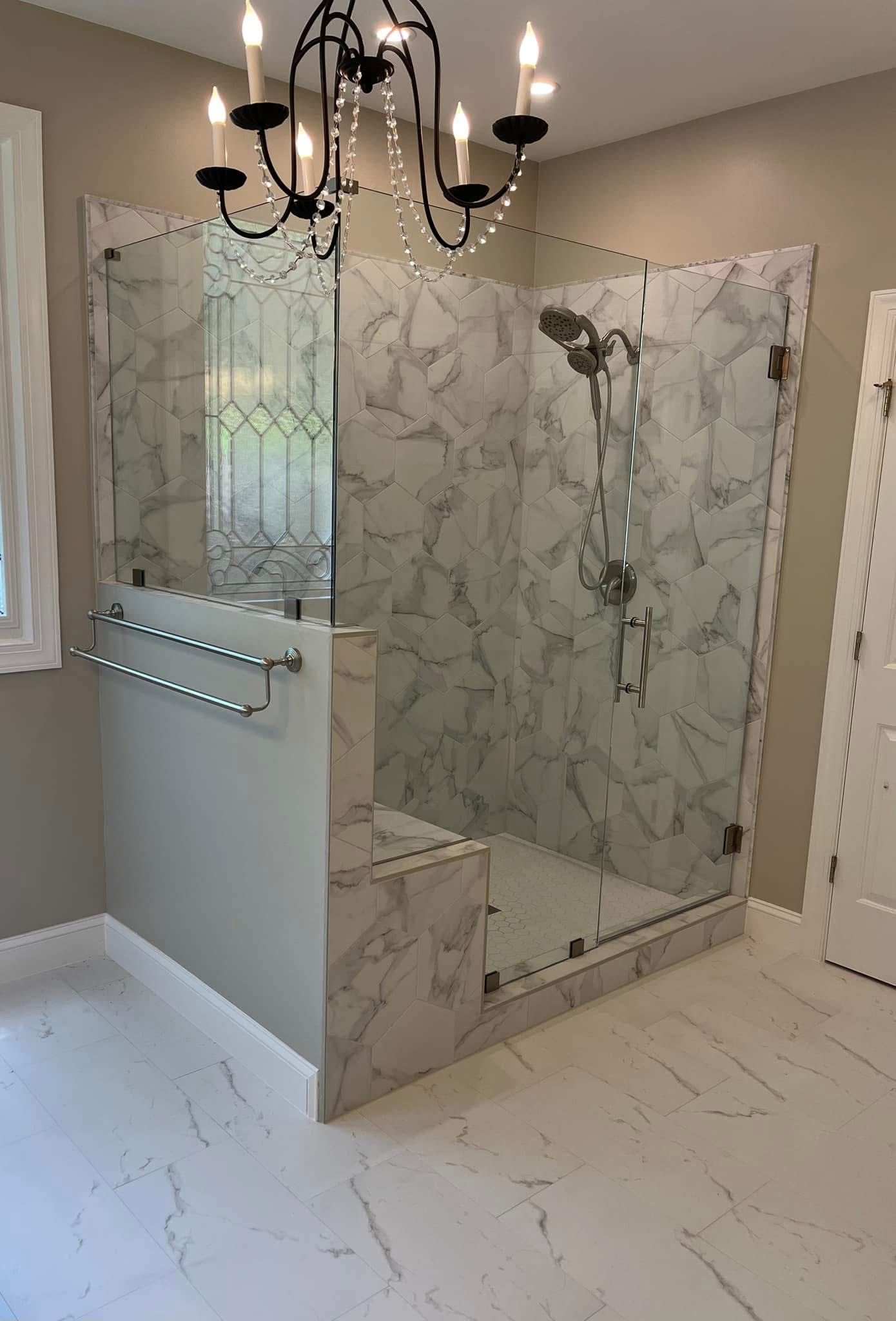 Elegant marble-tiled shower with glass doors, a bench, and a chandelier in a spacious bathroom.