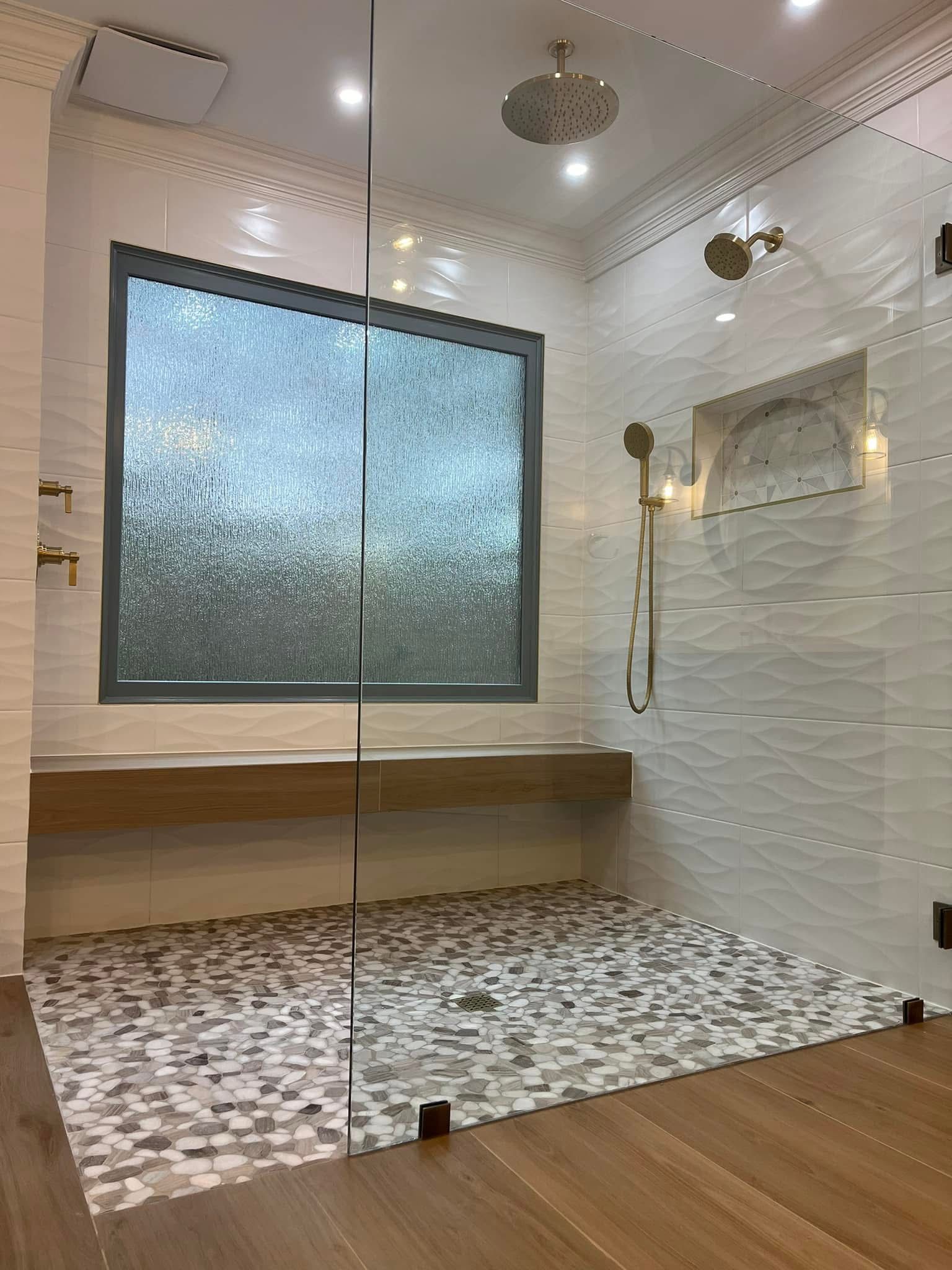 Modern shower with pebble floor, wood bench, and textured white tile walls. Glass enclosure.