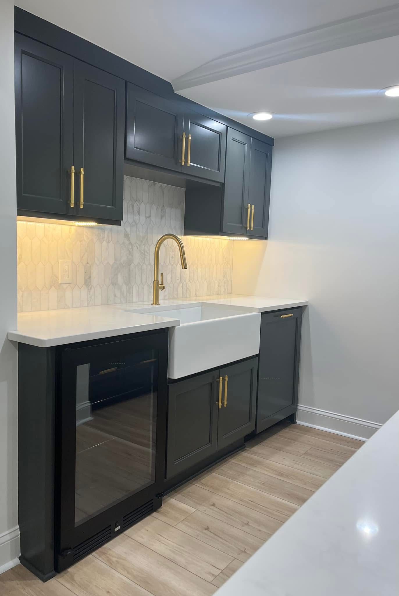 Dark grey kitchen cabinets with gold hardware, a white sink, countertop, and a wine fridge.