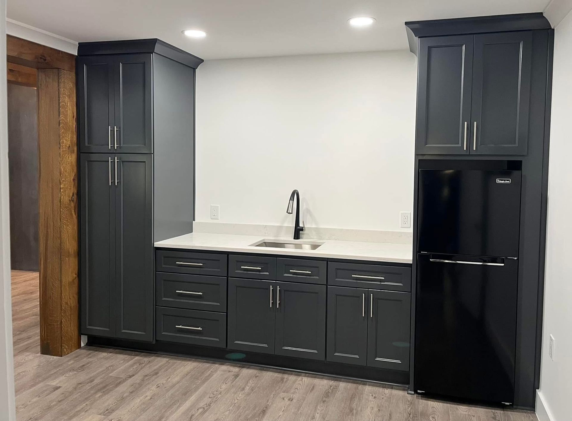 Dark gray cabinets with countertop, sink, and fridge in a room with light-colored flooring and white walls.