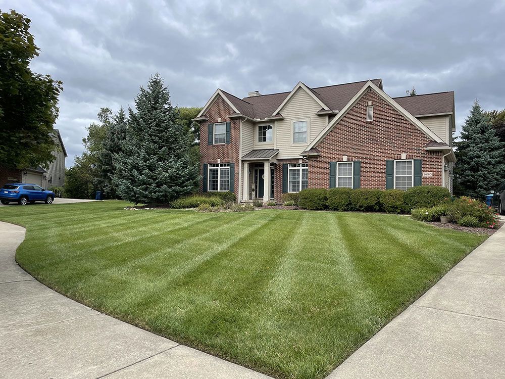 A two-story brick and stucco suburban house with a neatly mowed lawn, a paved driveway, and evergreen trees.