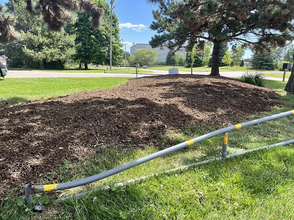 A large pile of dark brown wood mulch sits on a patch of grass in front of a tree, bordered by a metal safety rail.