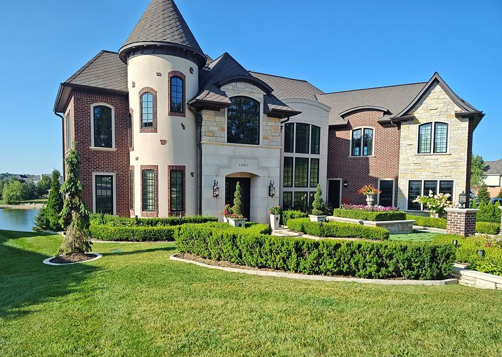 A large, two-story stone and brick house with a turret, manicured hedges, and a lawn beside a lake under a clear sky.