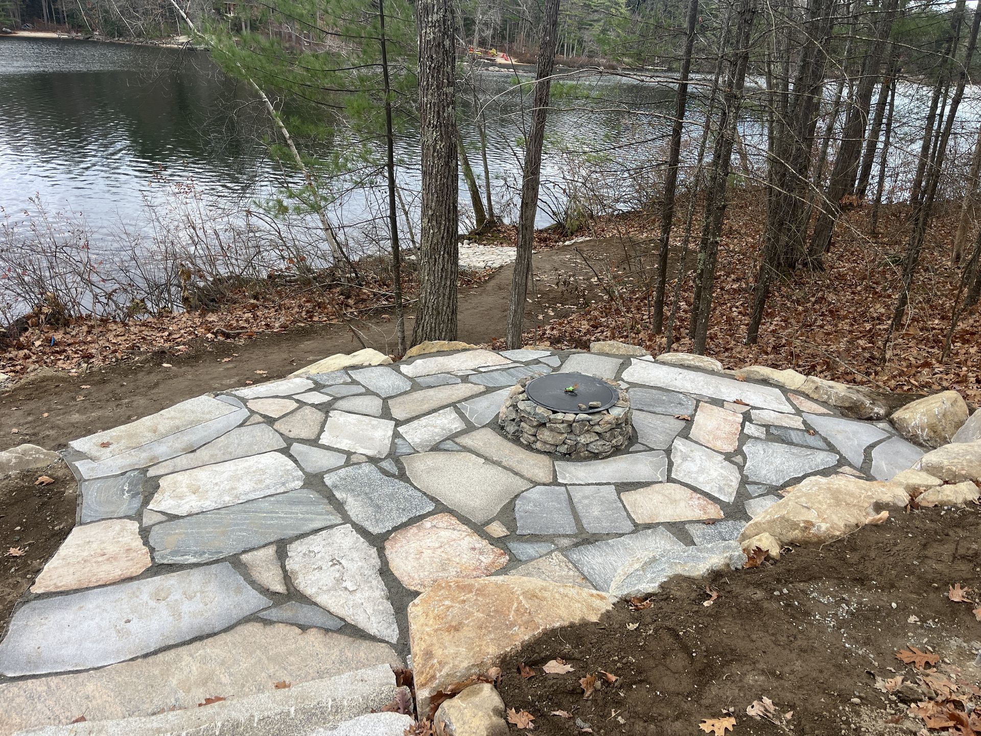 A stone patio with a fire pit and a lake in the background.
