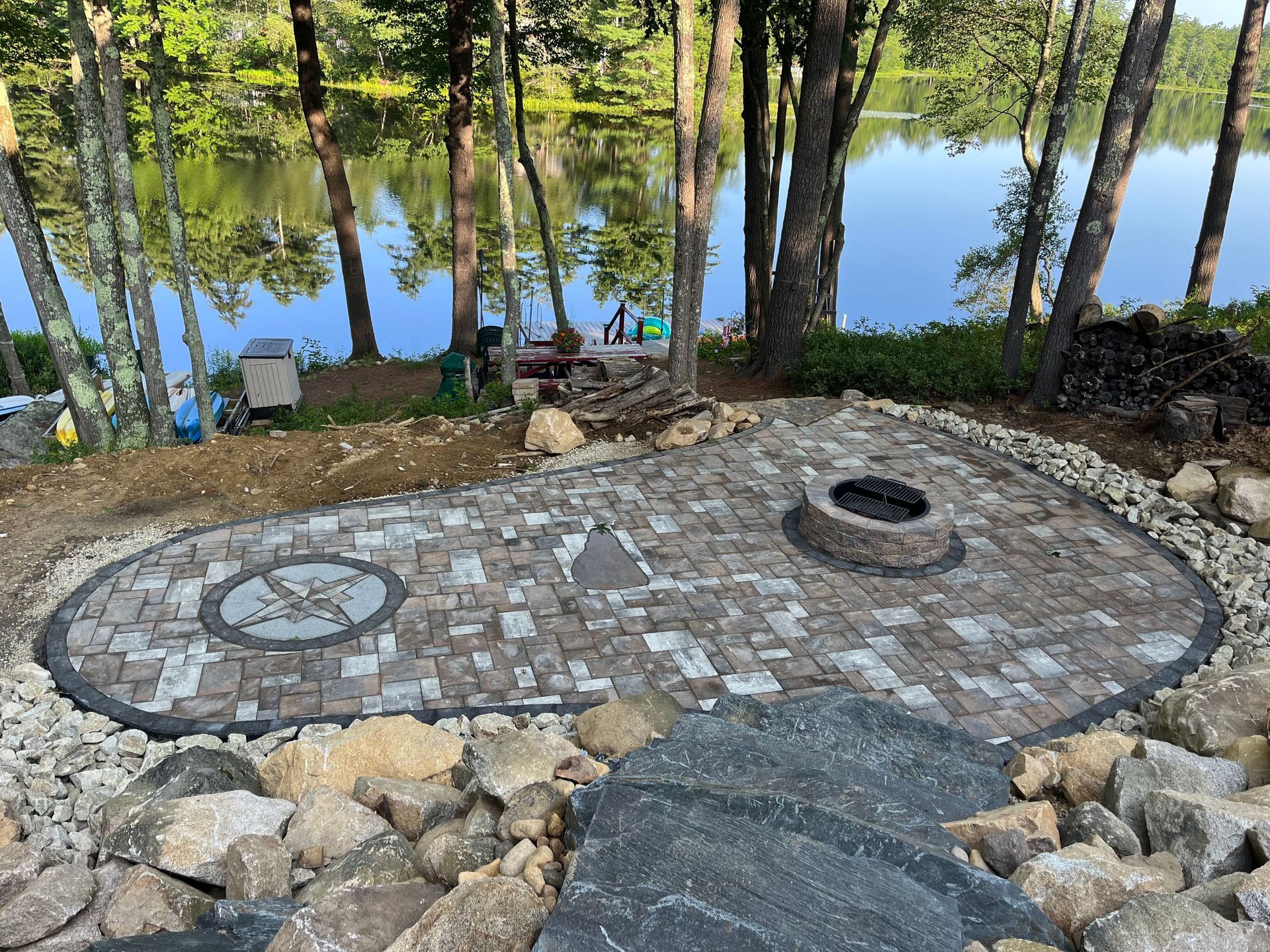 a fire pit in the middle of a patio next to a lake surrounded by trees