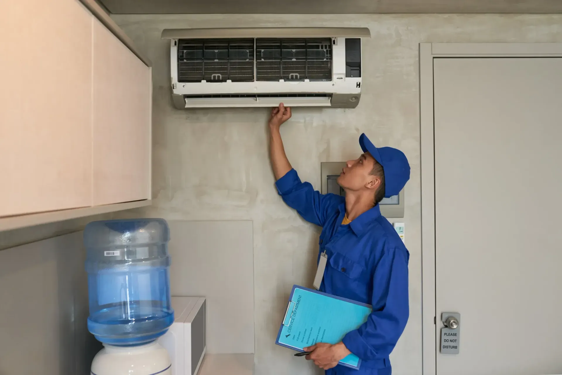 Technician in blue uniform inspecting a wall-mounted air conditioner next to a water cooler.