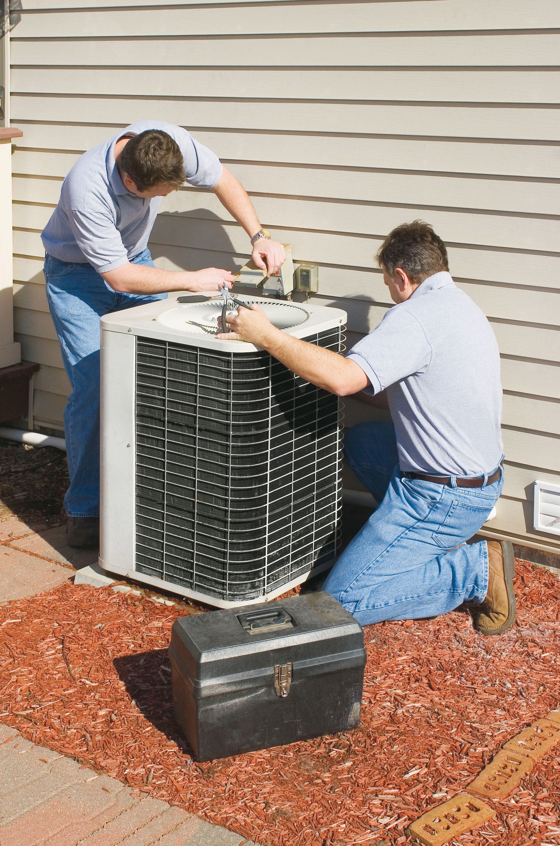 Two workers repairing an outdoor air conditioner unit beside a house, with a toolbox on the ground.