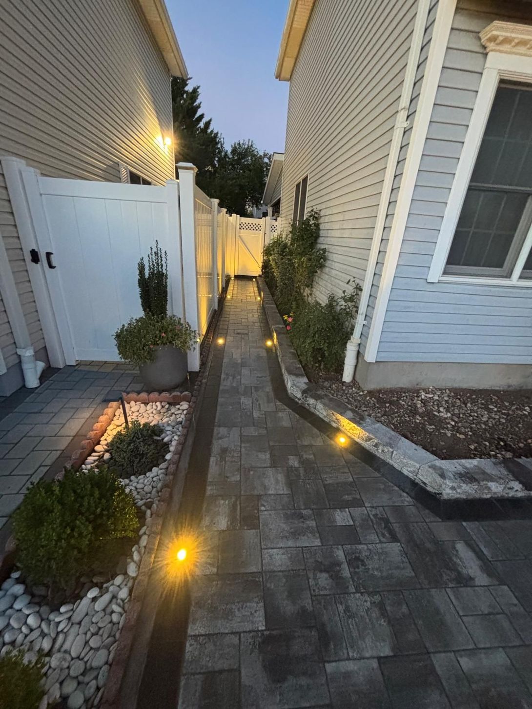 Narrow paved walkway between houses at dusk, lit by small ground lights and bordered by plants and a white fence.