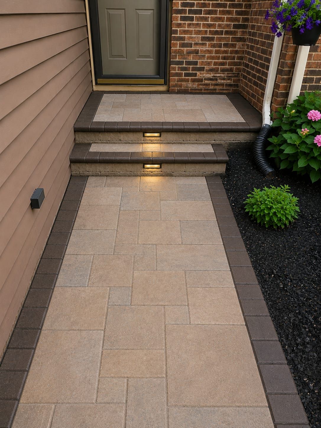 Front walkway with tan pavers, lit steps, brick and siding entry, and small landscaped shrubs.