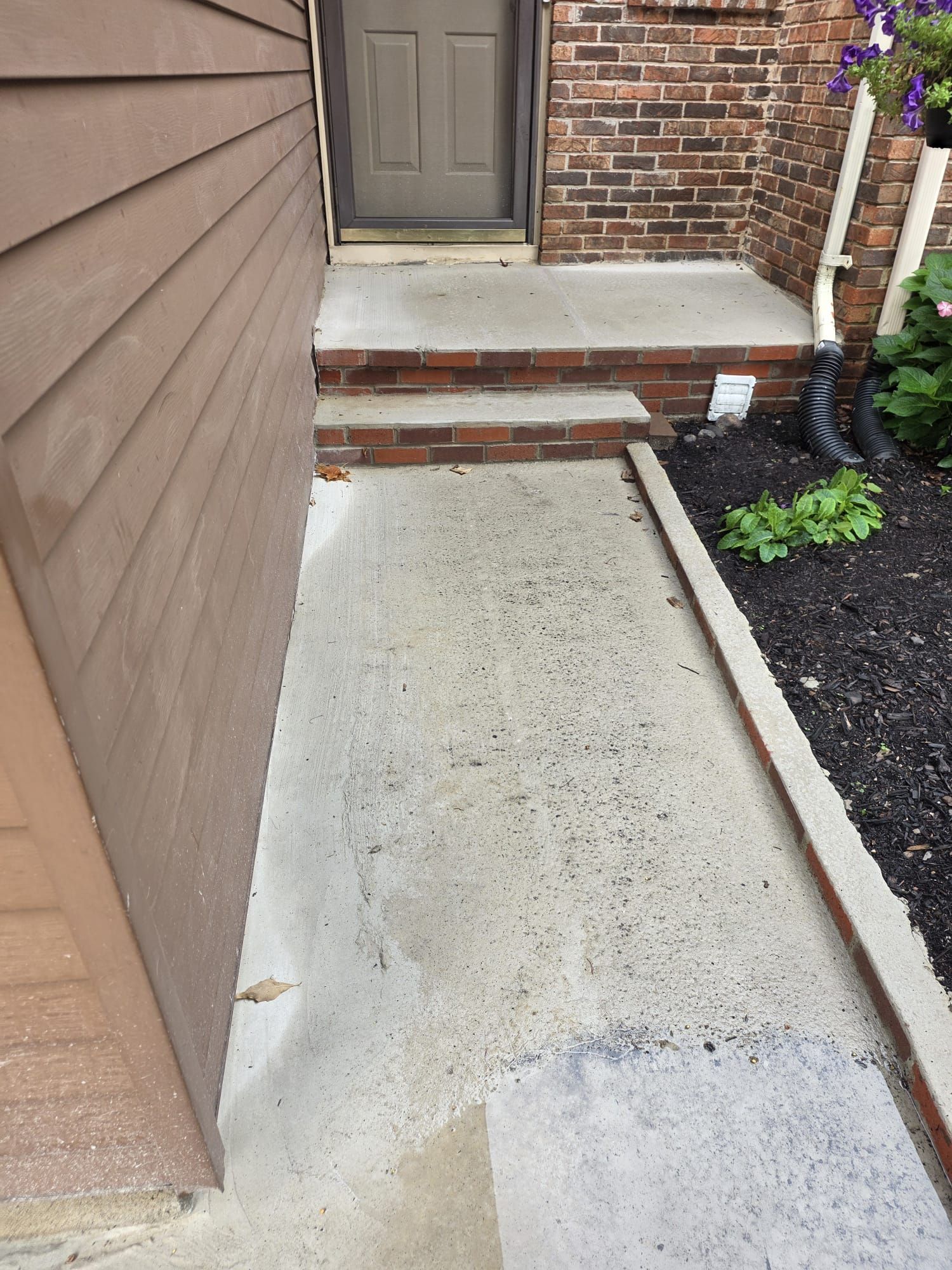 Concrete walkway to front steps and door beside brick house and mulch bed