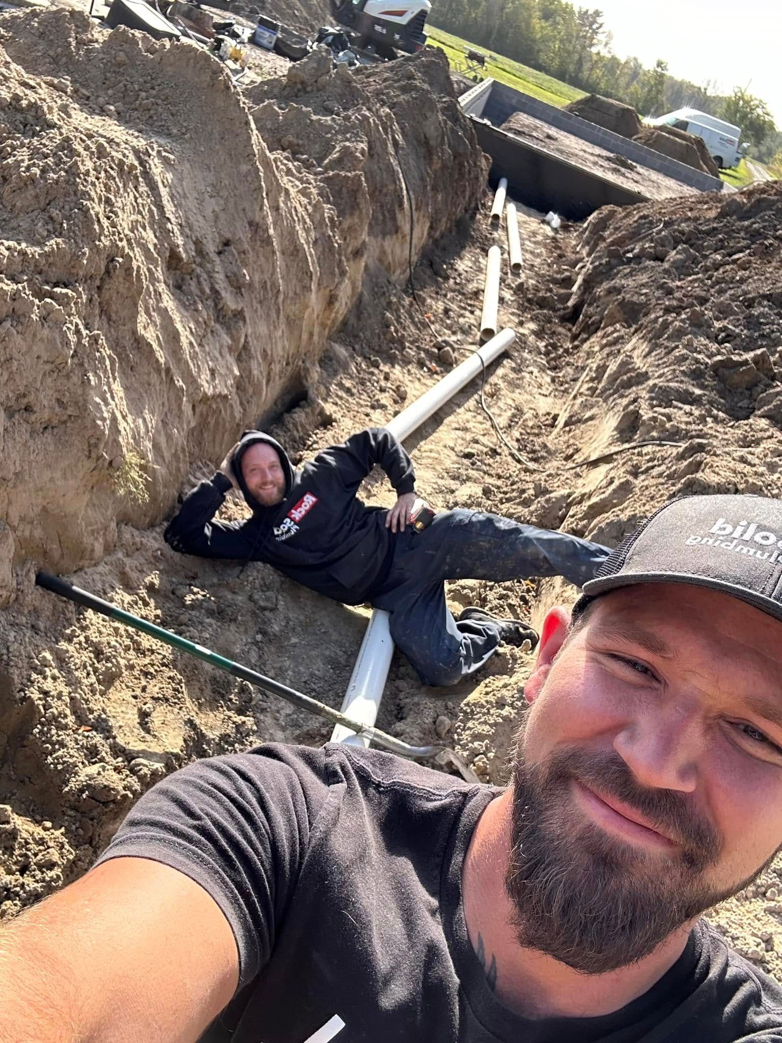 A man is taking a selfie while another man is laying in the dirt.