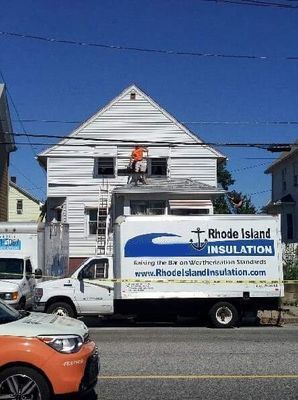 Rhode Island Insulation truck parked near a house with worker on roof, blue sky above.