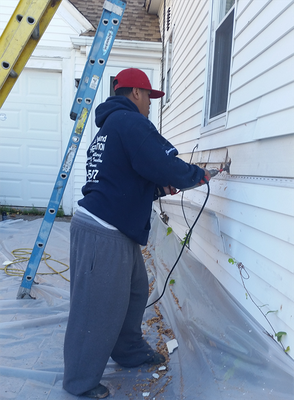 Person in blue hoodie using a tool on a house's siding. Ladder and plastic cover present.