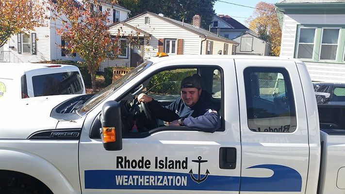 Man driving a white Rhode Island Weatherization truck on a sunny street.