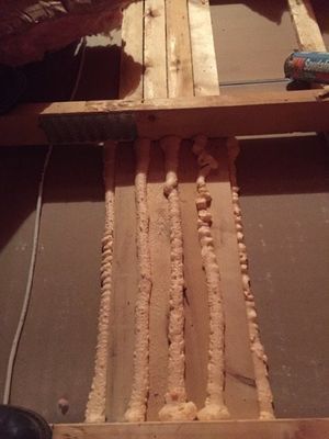 Wooden structural supports covered in expanding foam, between horizontal beams. Interior, dim lighting.