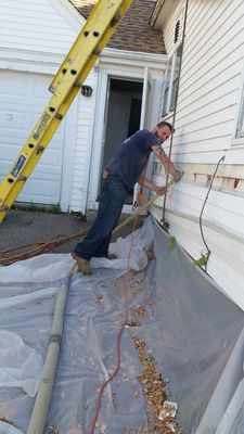 A man works on the side of a white house. He is standing on a plastic sheet. A ladder is propped against the house.