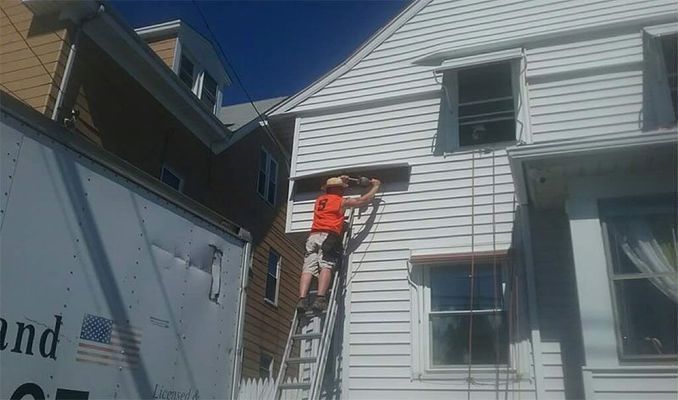 Person on a ladder, working on the white siding of a house, with a moving truck nearby.