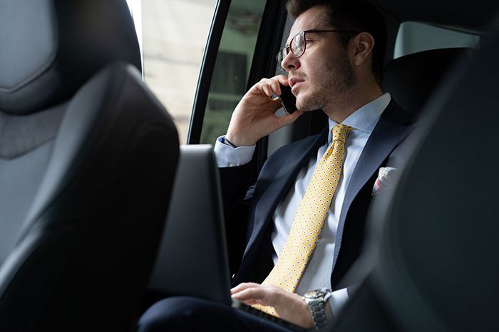 Man in suit on phone in car, working on laptop. He is wearing glasses and a yellow tie, looking out window.