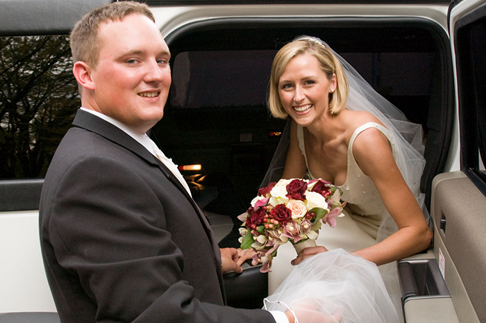 Groom helps bride in a wedding dress exit a white limousine. Both smile, holding a bouquet.