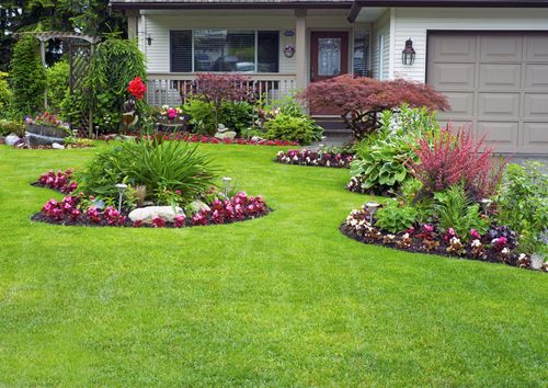 A residential front yard features a manicured green lawn with two rounded garden beds planted with flowers and shrubs.