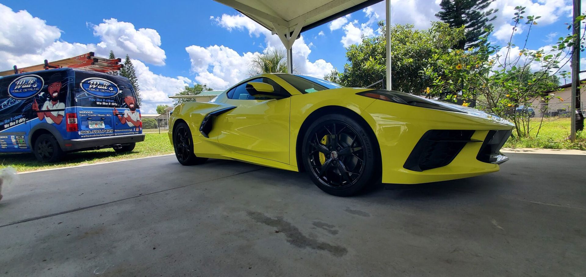 Yellow sports car parked under a canopy next to a blue van; sunny day.