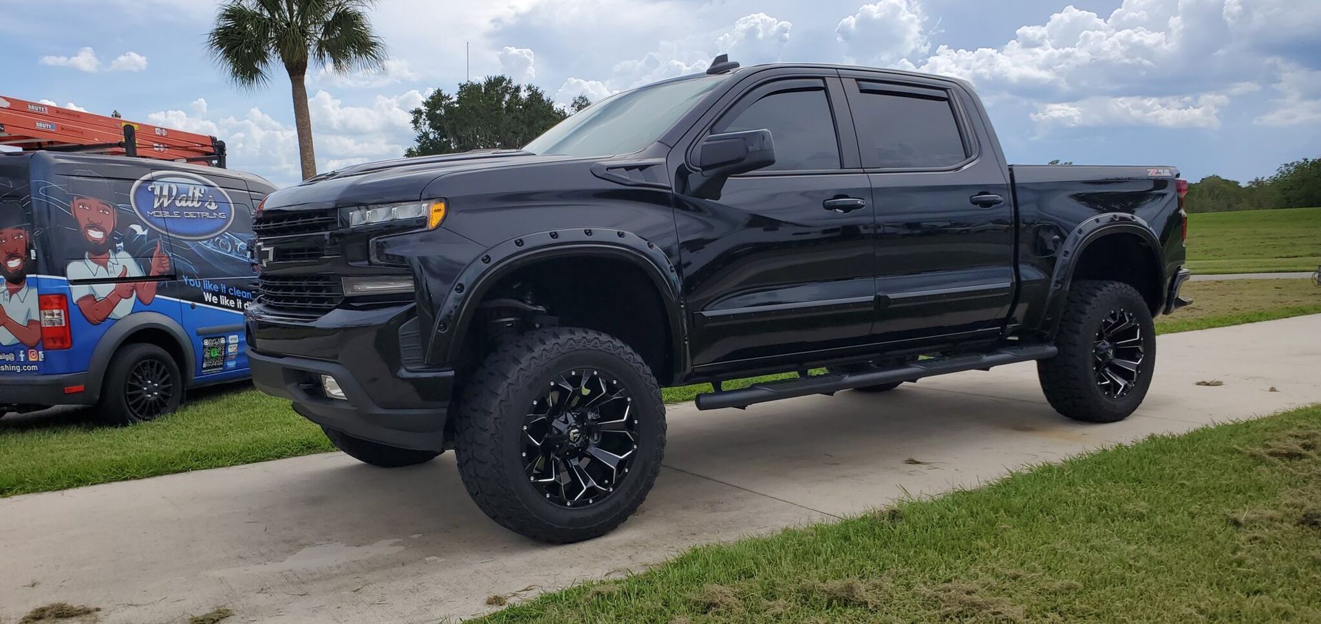 Black truck with large wheels parked on a concrete path, with a van parked in the background.