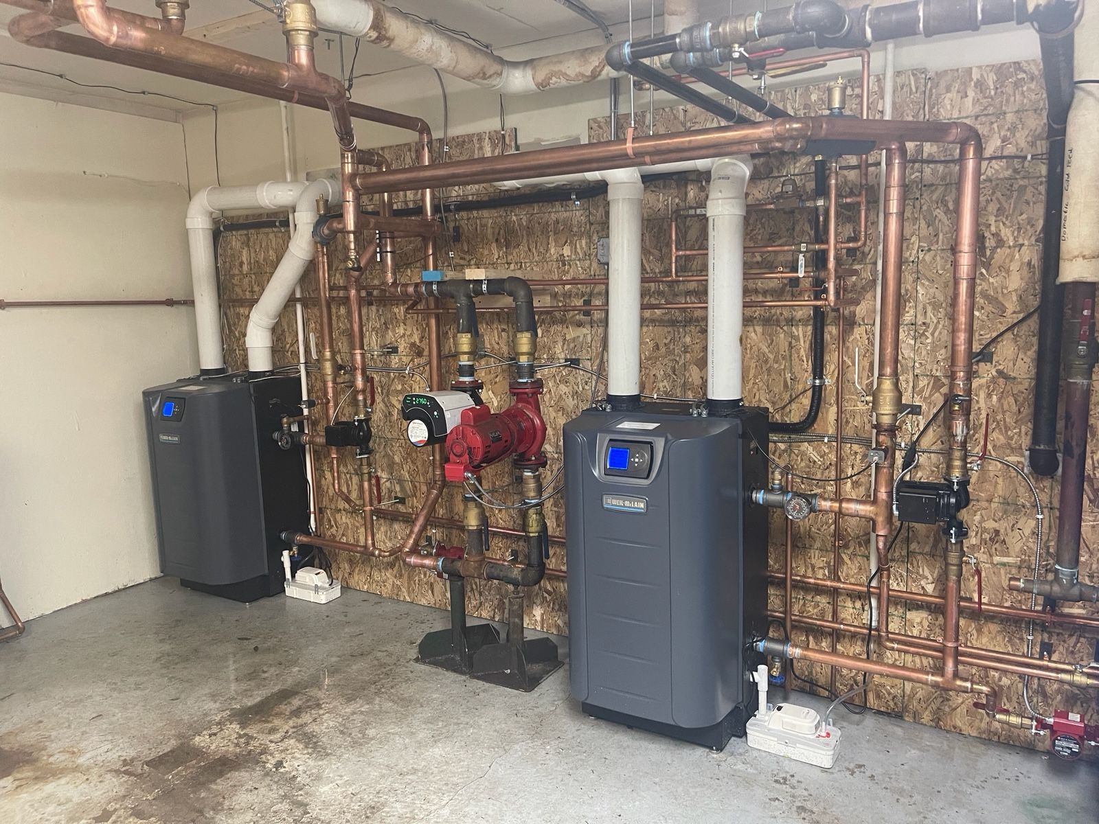 Two boilers are sitting next to each other in a room with pipes.