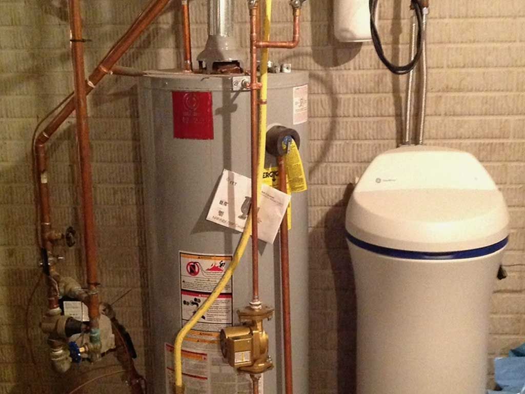 Water heater and plumbing setup beside a white water softener in a basement utility room