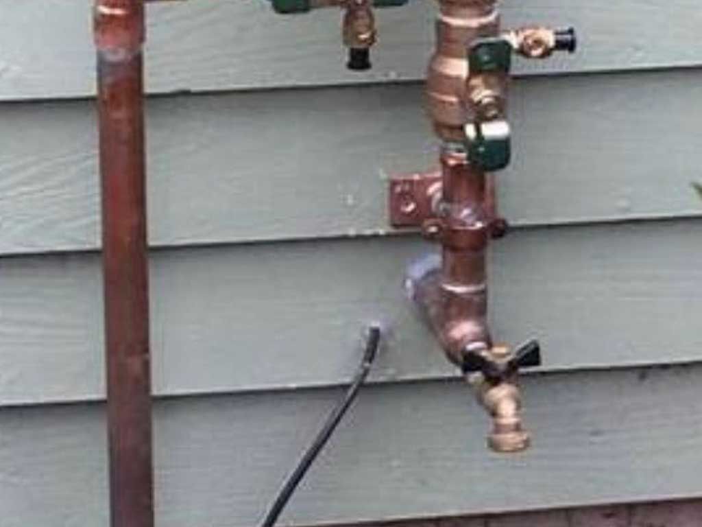 Rusty outdoor plumbing pipes and valves mounted on gray house siding, with a black cable below.