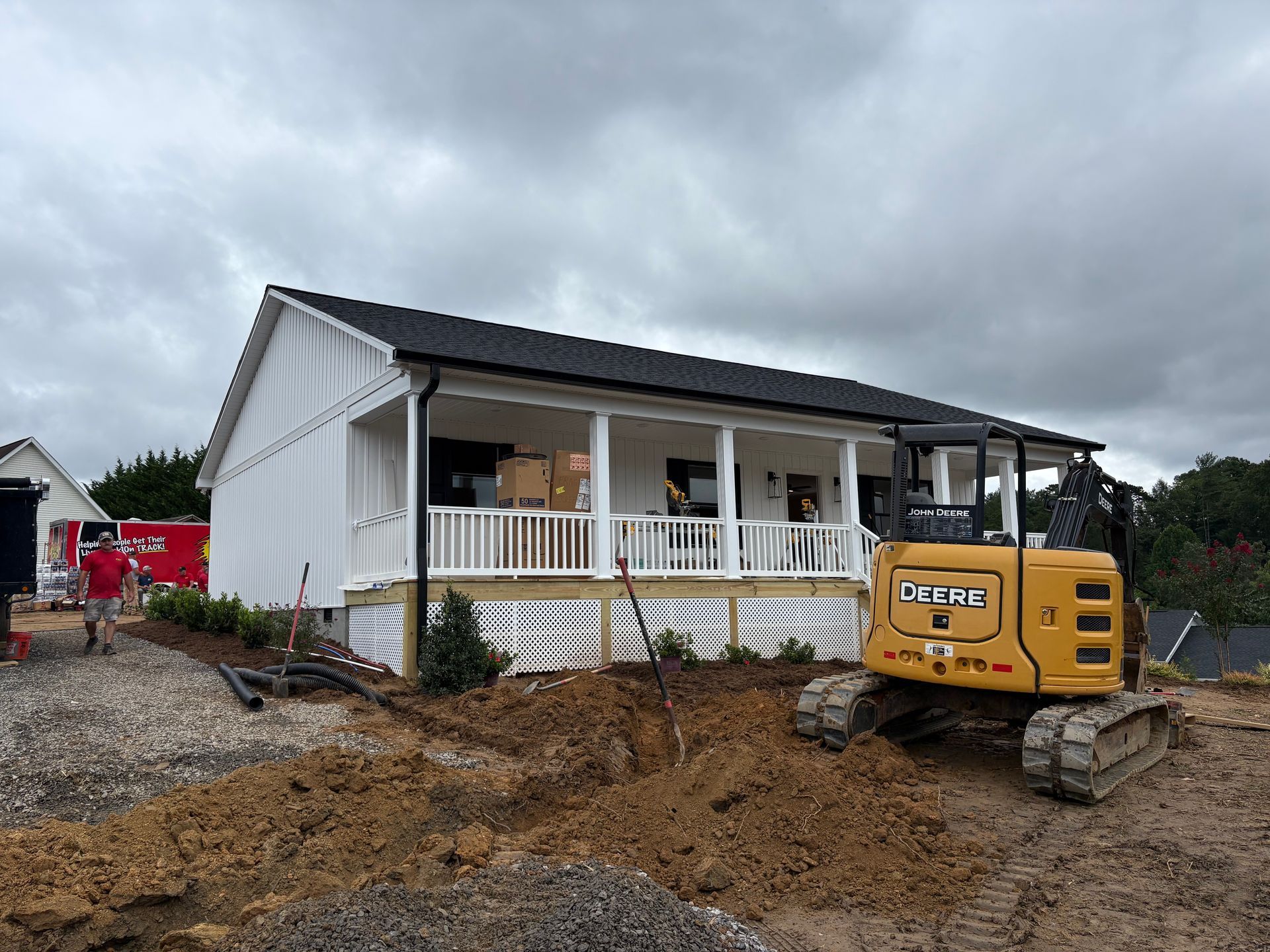 A white house with a porch is under construction; a yellow excavator sits in the foreground, cloudy sky.