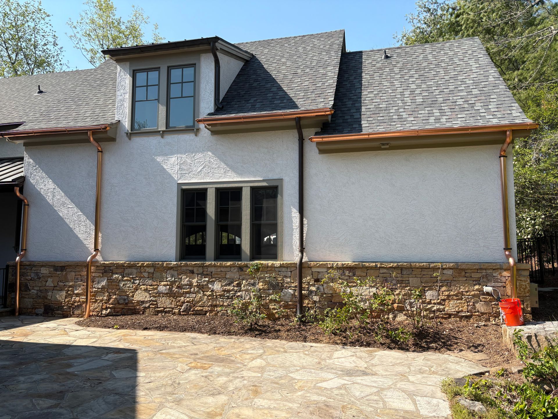 Tan stucco house with copper gutters, stone base, and landscaping, under a blue sky.
