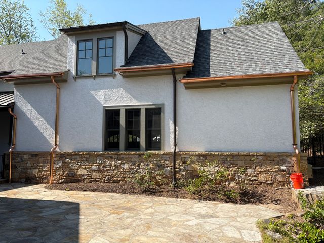 Tan stucco house with copper gutters, stone base, and landscaping, under a blue sky.