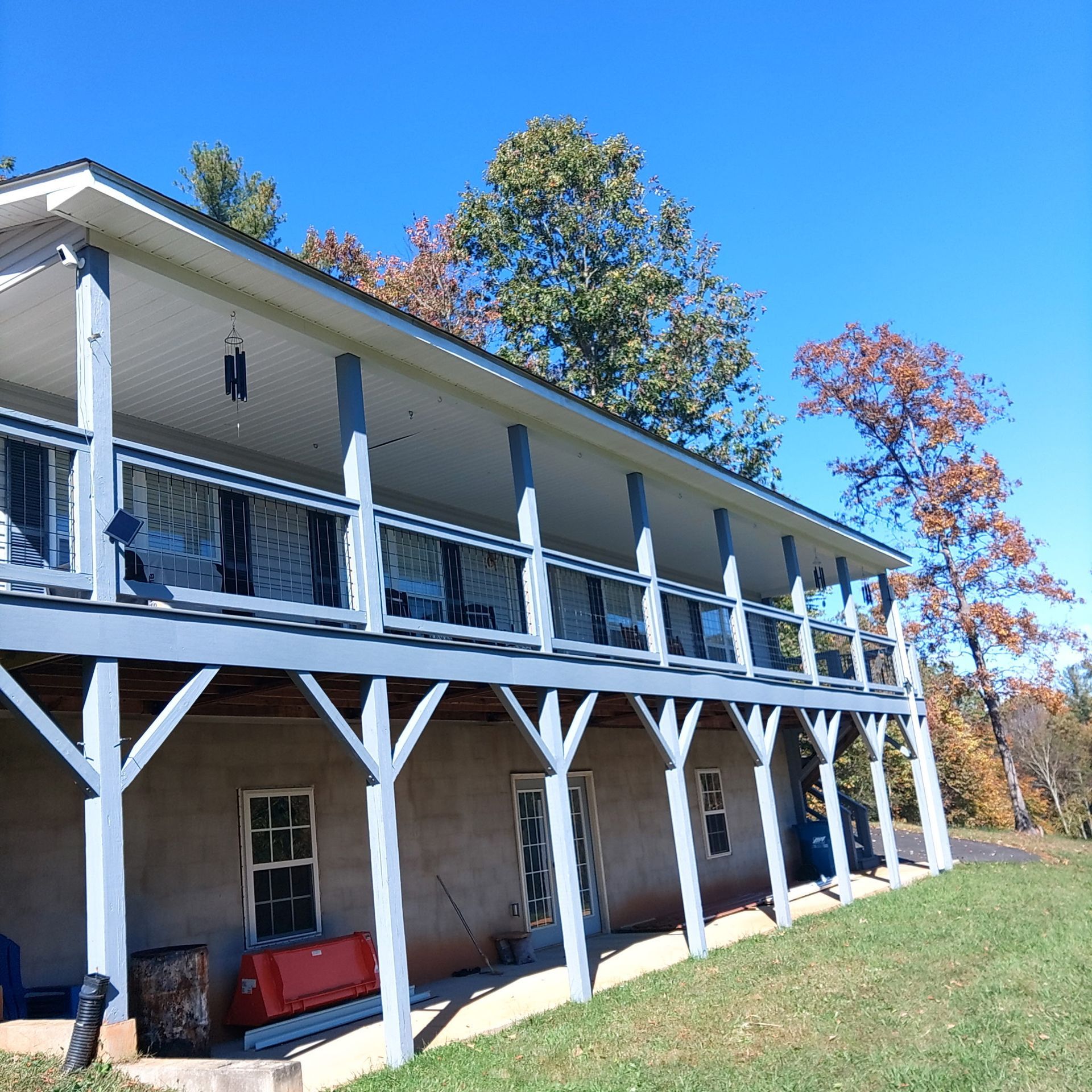 Two-story house with a large porch supported by white and gray beams. Trees and clear blue sky in the background.