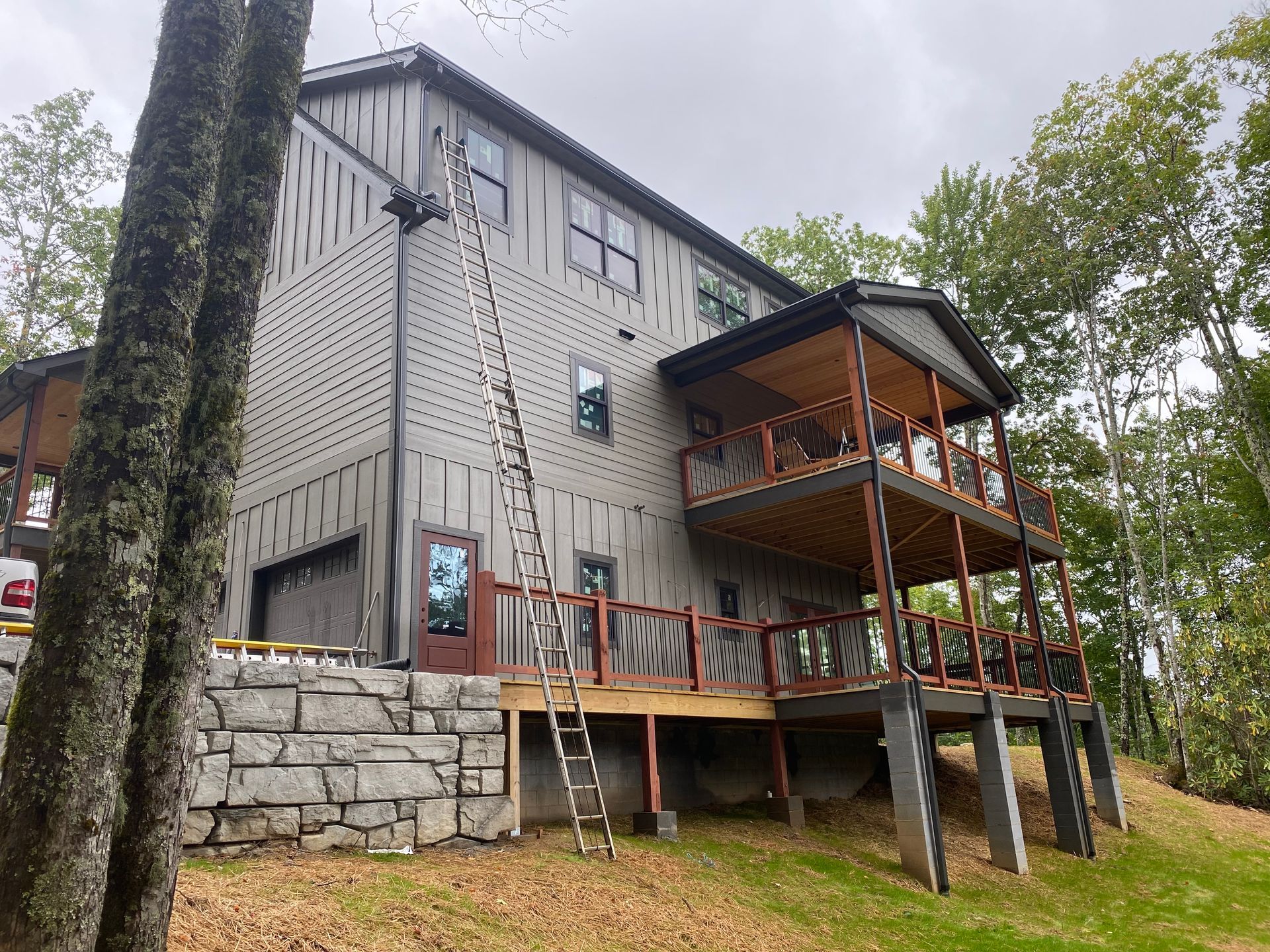 Two-story gray house with a wooden deck and retaining wall. A ladder is leaning against the side.