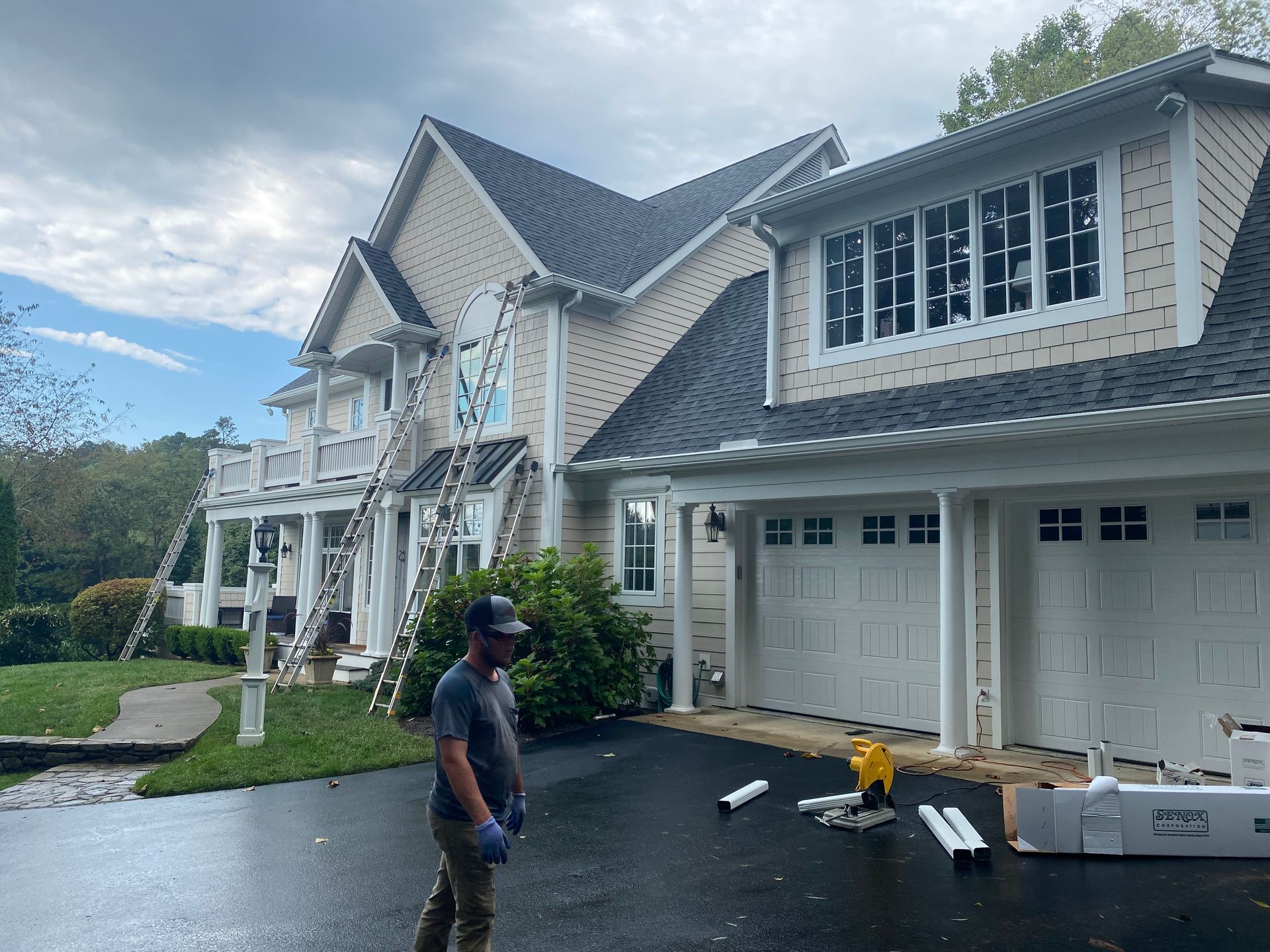 Exterior house with siding work in progress; worker standing in driveway, ladders on house.