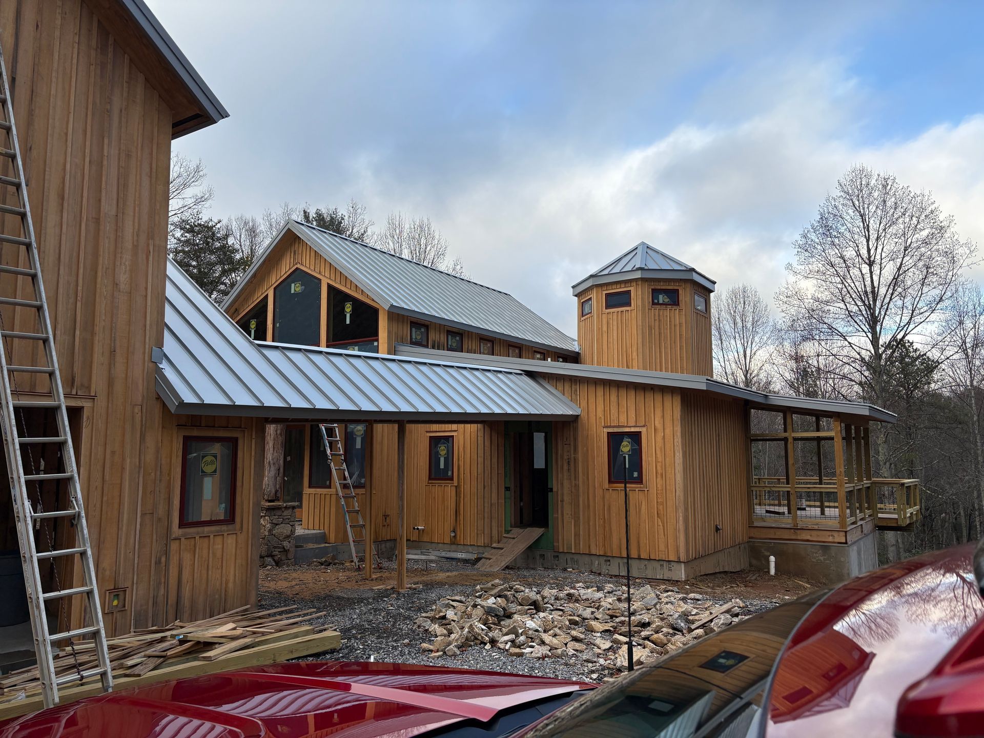 Wooden house under construction with a metal roof. A ladder leans against the siding, car in foreground.