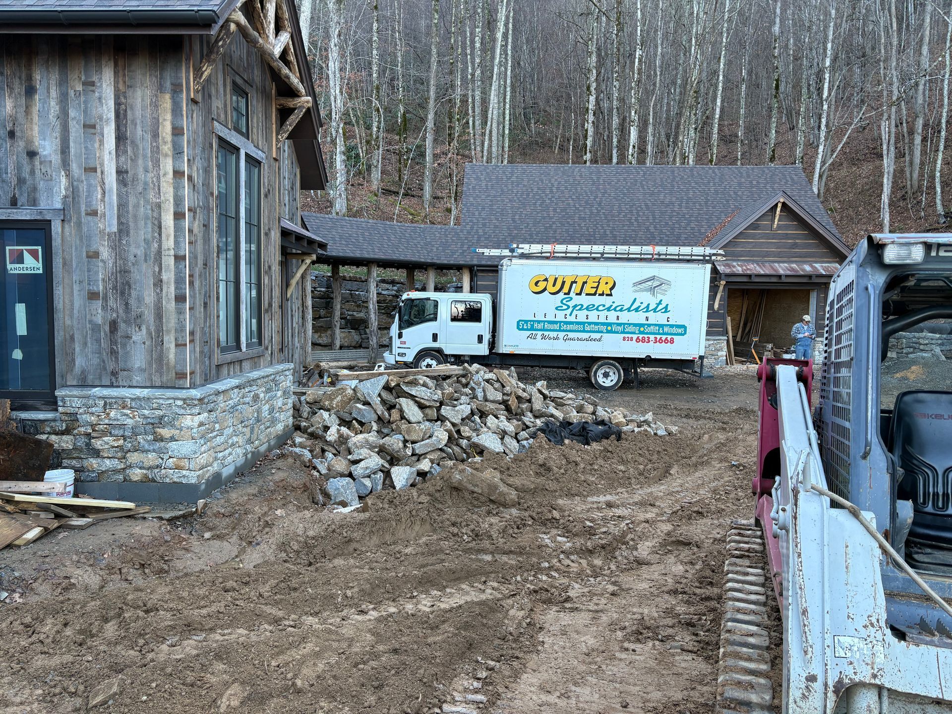 Construction site: wooden house, stone foundation, white truck, rocks, and skid steer.