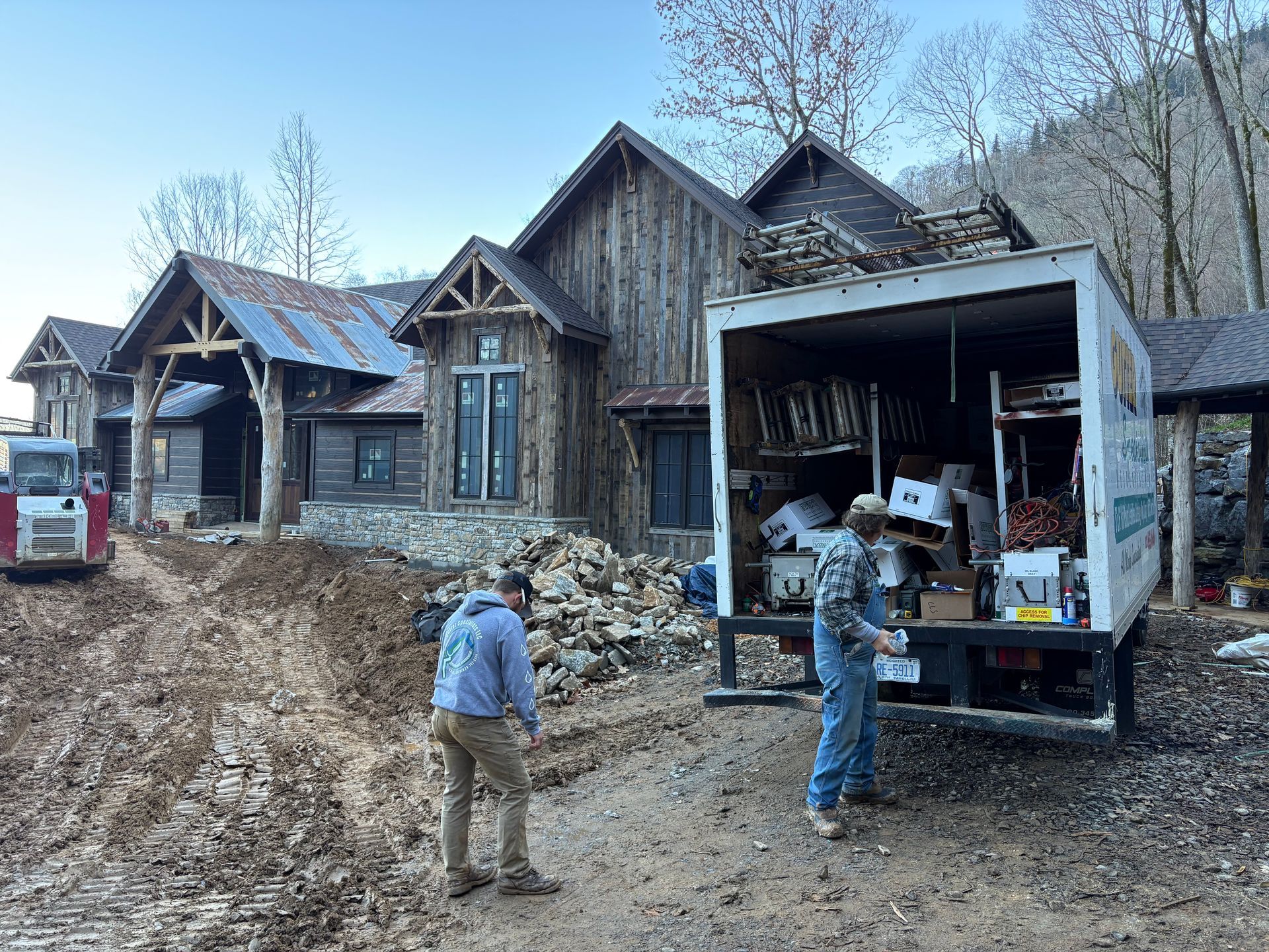 Men unloading materials from a truck in front of a rustic house under construction.