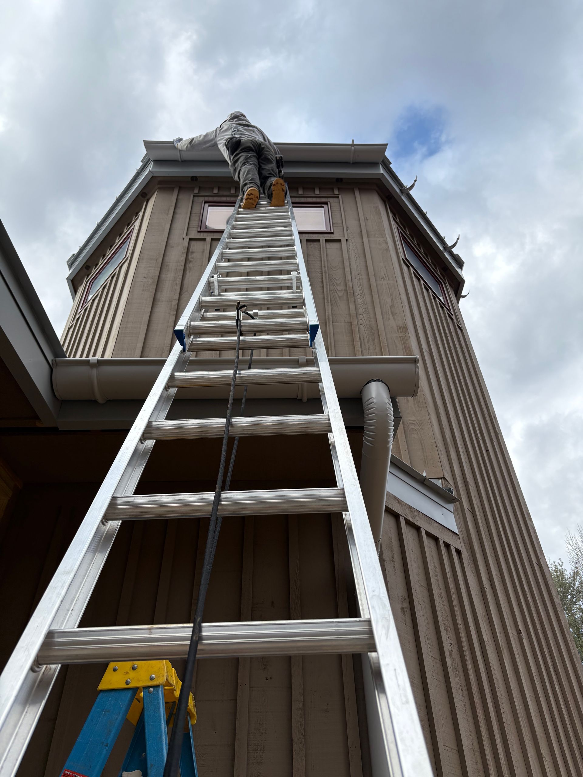 Person on a ladder, cleaning the gutters of a tall, light brown building against a cloudy sky.