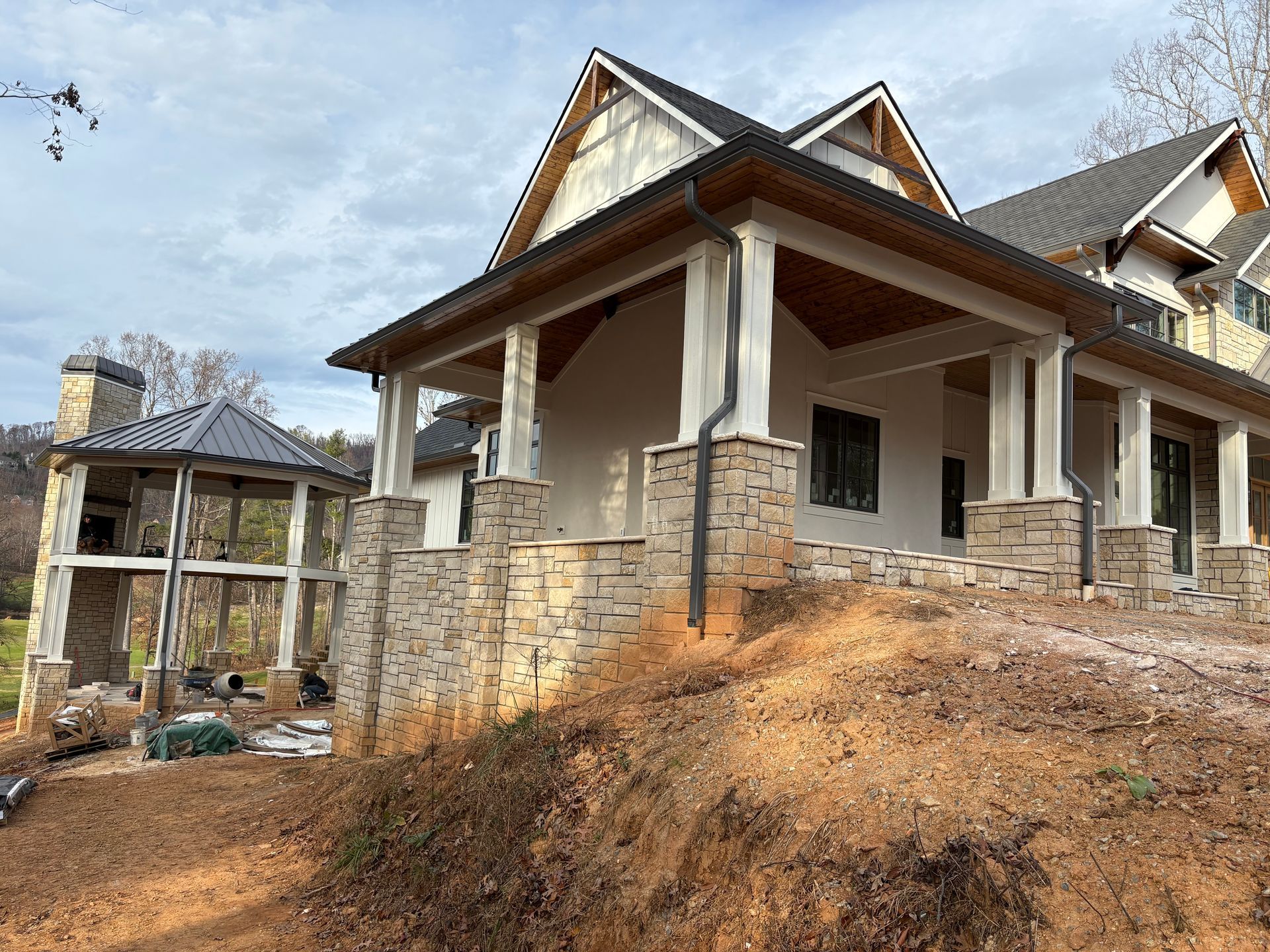 Stone and white house with covered porch on a hillside, and a detached structure with a fireplace.