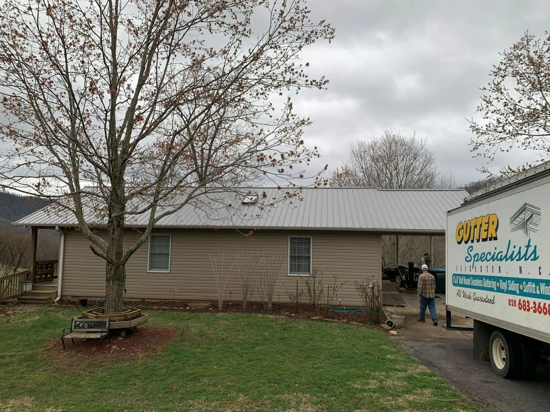 House with metal roof, truck, tree, and person standing. Cloudy sky.