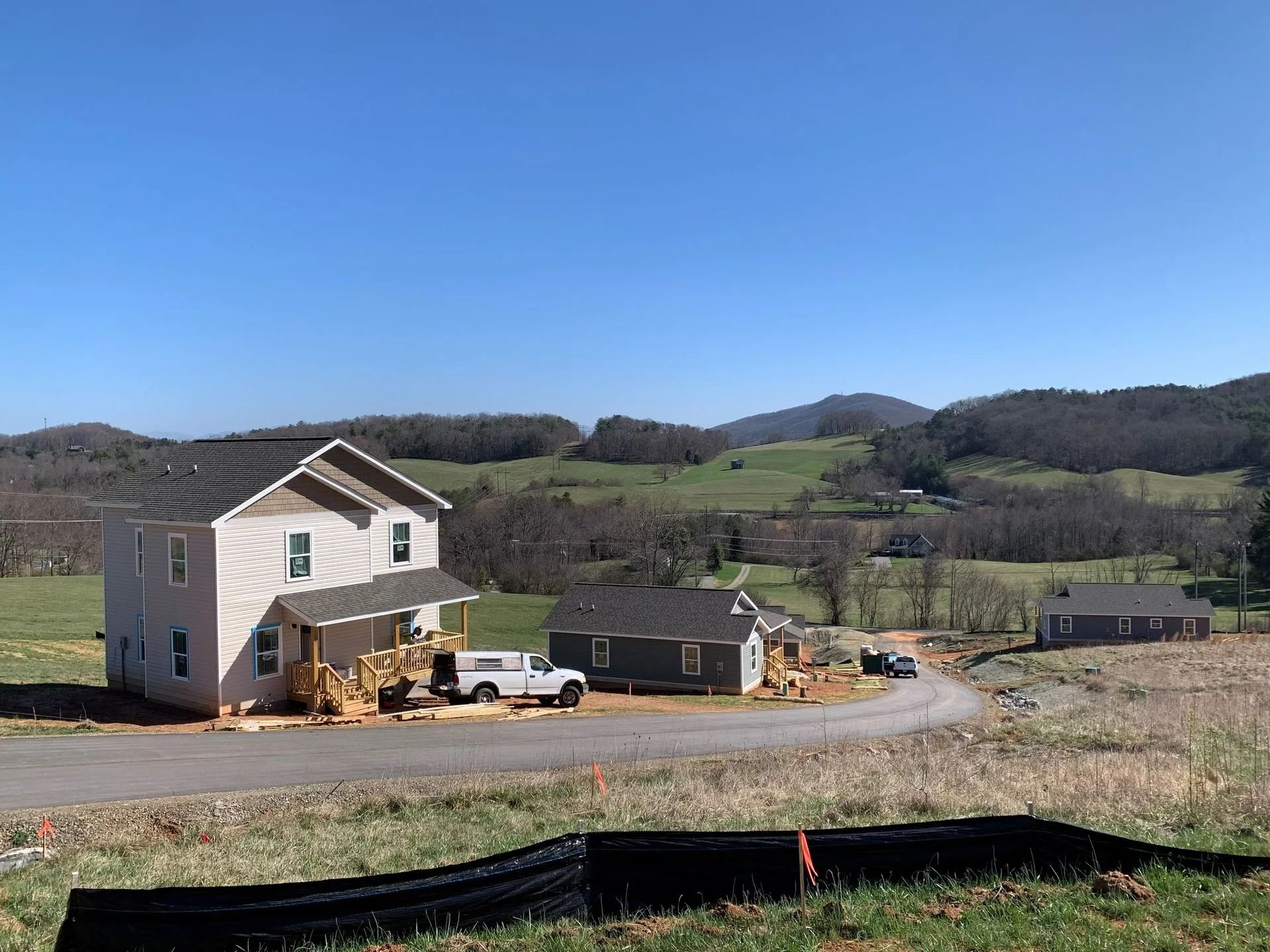 Houses under construction on a curved road, green hills and mountains in the background. Blue sky overhead.