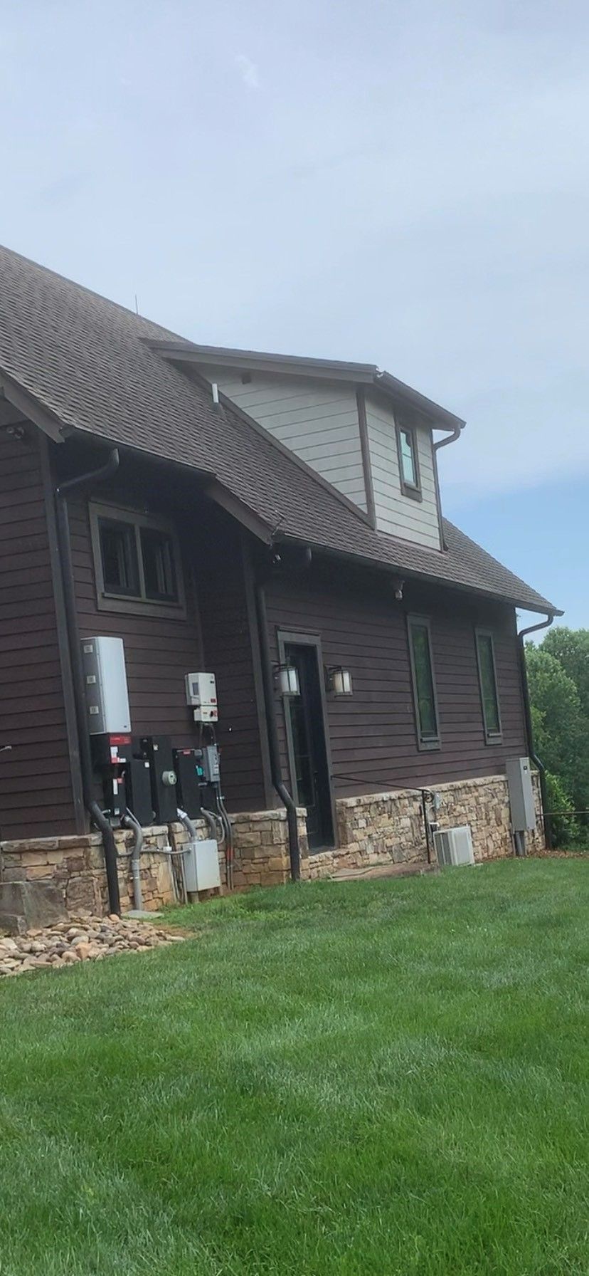 Brown house with a stone foundation and green lawn, with electrical equipment visible.