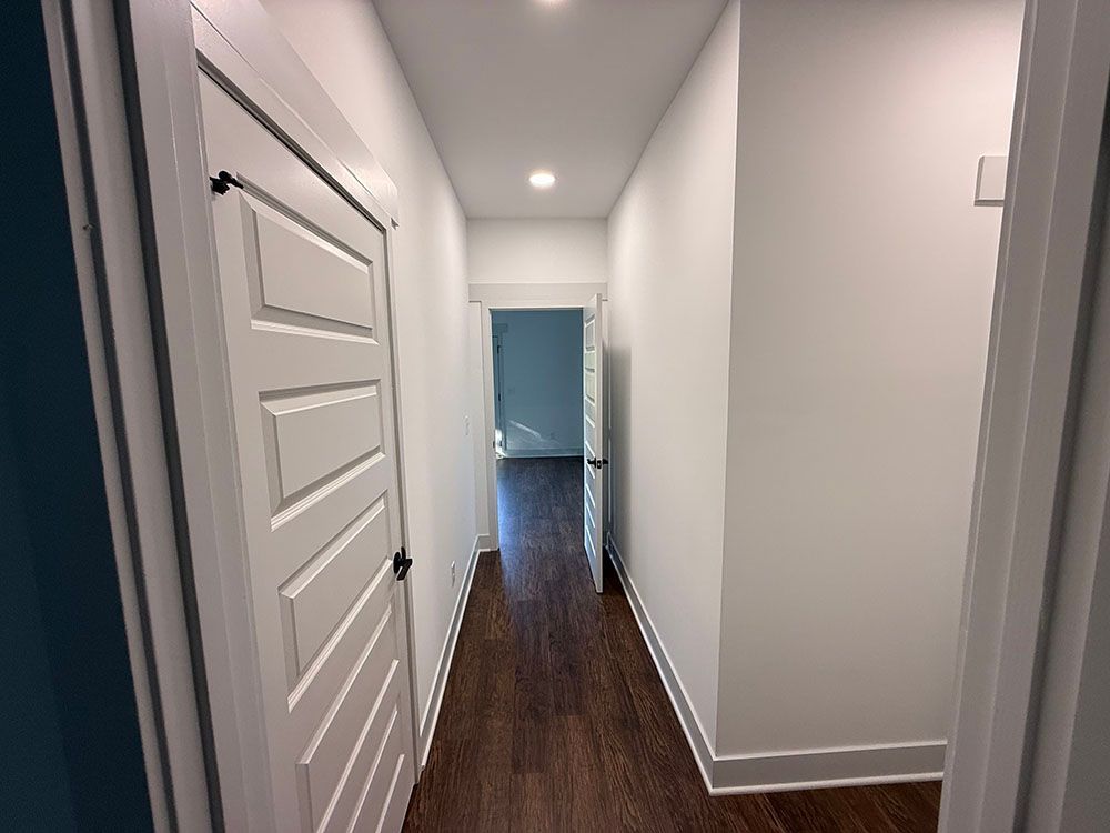 Narrow hallway with white walls, wood-look floor, and doors on the left and center, leading to a room.