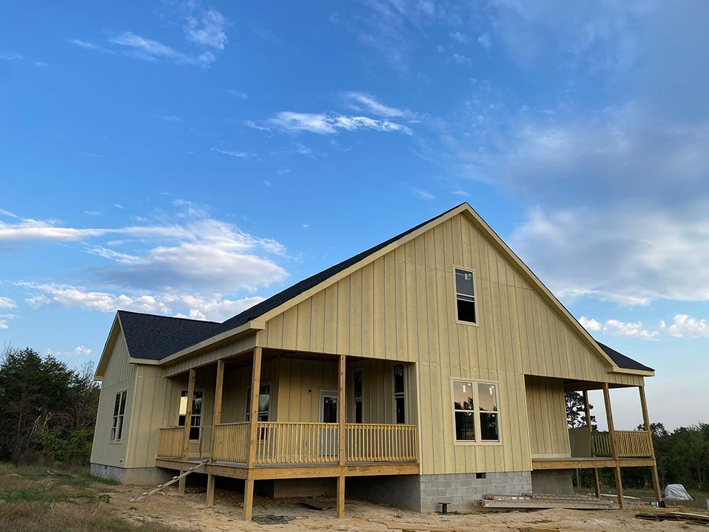 New construction house with light-colored siding, porch, and dark roof against a blue sky with clouds.